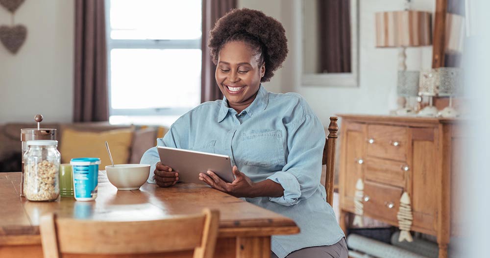 Middle-aged African American women smiling at her tablet while sitting at a wooden dining table.