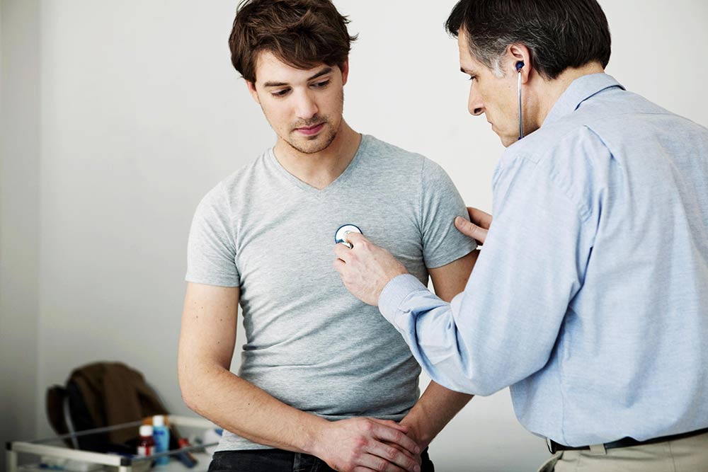 Doctor using stethoscope to check the heartbeat of patient sitting on an exam room table. 