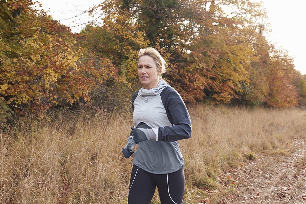 Middle-aged women jogging in a frosted farm field with fall colored trees behind her. 