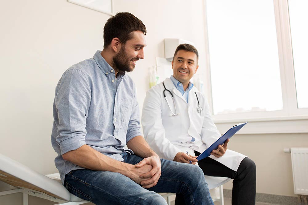 Doctor and patient sitting side by side on an exam room table, smiling while looking at a clipboard.
